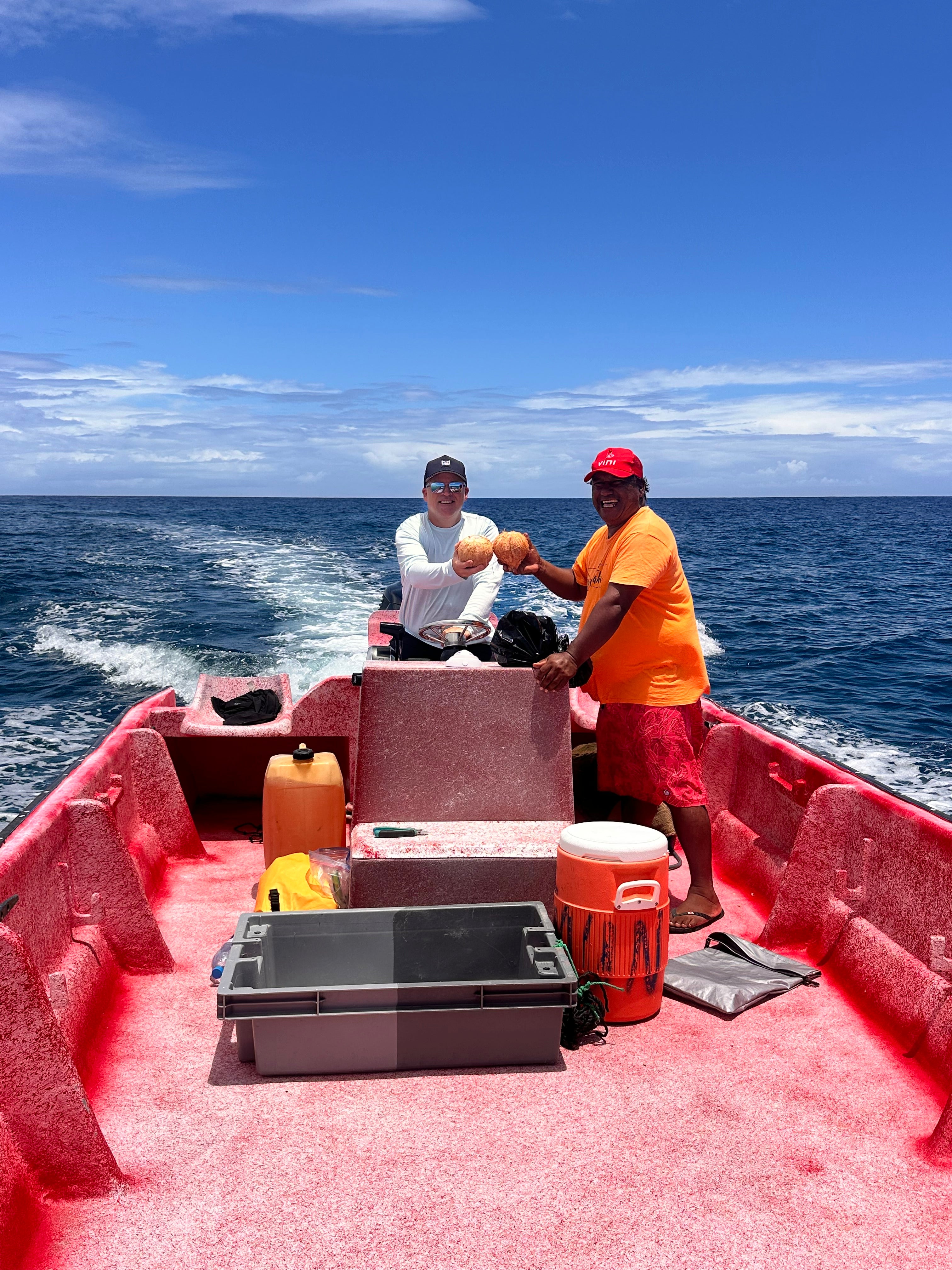 Two men on a boat in the middle of the ocean, smiling and doing a cheers with cracked open coconuts.