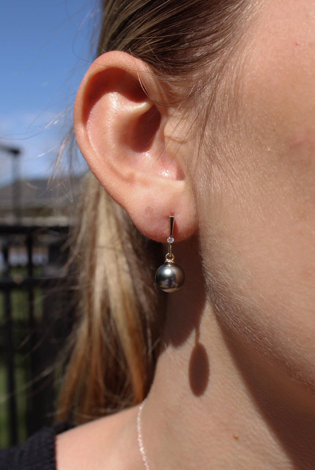 Close-up of a person wearing a yellow gold and lab grown diamond earring featuring a black pearl with a blurred outdoor background