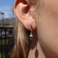 Close-up of a person wearing a yellow gold and lab grown diamond earring featuring a black pearl with a blurred outdoor background